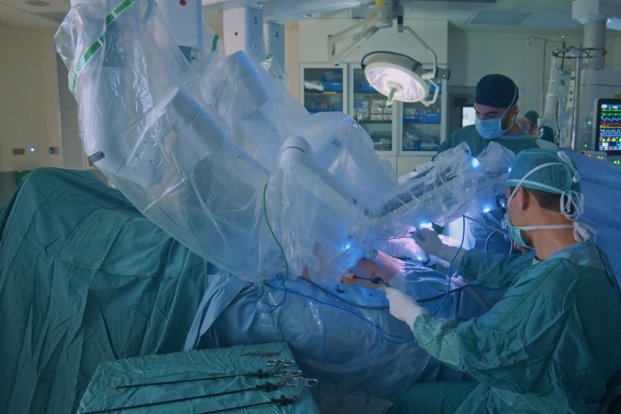 A imagem é uma cirurgia robótica na coluna, possui médicos vestido de azul operando uma pessoa com auxílio de robôs.